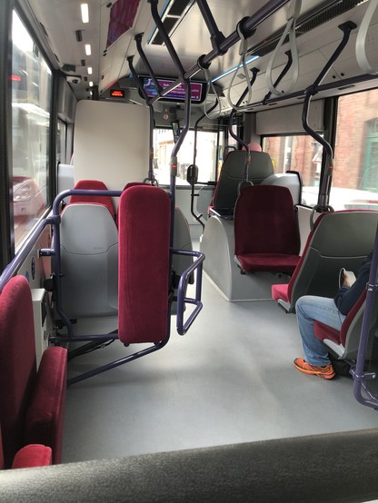 Vision de la partie avant de l'intérieur du bus. Les sièges assis sont plus larges, il y a des strapontins et des lanières sur la barre aérienne